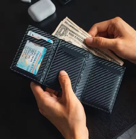 Person holding a carbon fiber wallet with money and an ID card on a dark surface